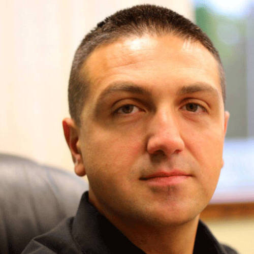 A man with short dark hair sitting in an office chair, wearing a black collared shirt, looking at the camera with a neutral expression.